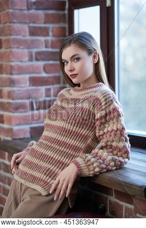 Attractive girl in stylish casial clothes stands by the window leaning on a windowsill in a loft room and slightly smiles. Happy and successful people. 