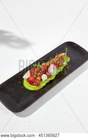 Modern food - tuna tartare on sliced avocado in black plate. Avocado starter with tuna and radish in asian style. Contemporary asian dish. Healthy food with avo and marinated fish on white background