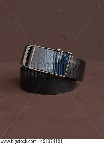 Rolled-up Black Fashionable Men's Leather Belt With Automatic Buckle On Brown Background. Male Acces