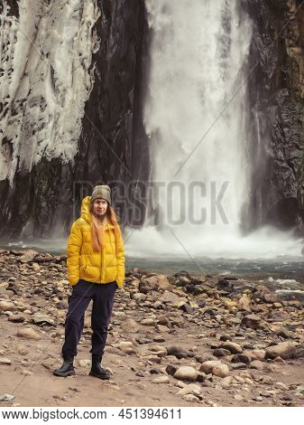 The Girl Stands Against The Backdrop Of A Large Powerful Waterfall Flowing From An Icy Rock. Soft Fo