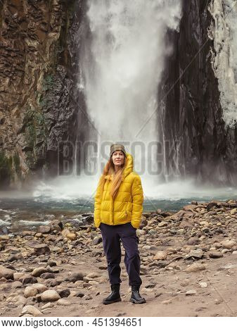 A Woman Stands On The River Bank Against The Backdrop Of A Stormy Waterfall Flowing Down From The Ro