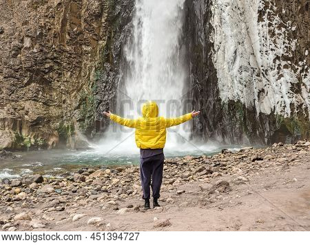 A Woman In A Yellow Jacket Stands With Her Arms Outstretched Looking At A Stormy Icy Waterfall Flowi