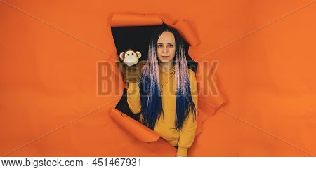 Young Woman With Soft Puppet Toy On Hand Looking Out Of Hole Of Orange Background. Pretty Female Wit