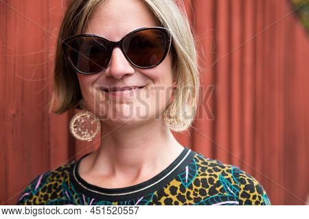 Portrait Of An Attractive Thrity Year Old Woman With Sunglasses Standing Against A Typical Falun Red