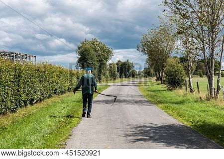Bullingen, Walloon Regio - East Belgium - 08 27 2019 An Old Farmer Carrying A Scythe, Looking Like T