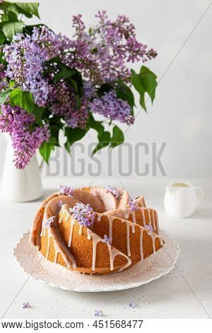 Classic Vanilla Bundt Cake With Sugar Glaze And Lilac Flowers On White Background. Spring Compositio