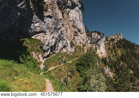 Two Mountaineers Hiking On A Diagonal Path Under A Stone Wall.