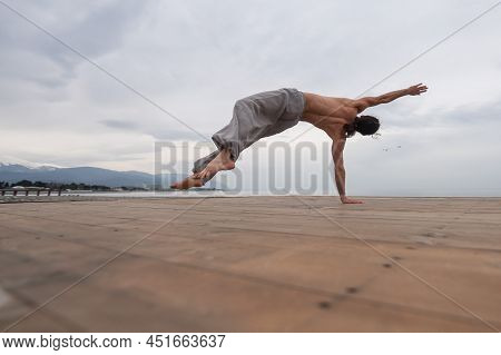 Shirtless Caucasian Man Doing Backflip On Pebble Beach.