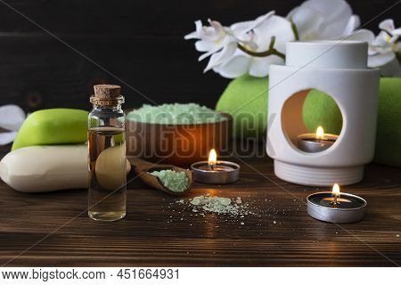 Body Oil, Tea Candles And Bath Soap On A Dark Brown Wooden Table. Green Bath Towel And Orchid Sprig 