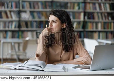 Thoughtful Girl Studying In Library Seated At Table With Textbooks