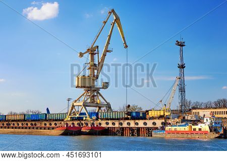 River Port Crane Loading Open-top Gondola Cars On Sunny Day. Empty River Drag Boats Or Barges Moored