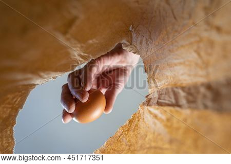 A Hand Places A Chicken Egg In A Paper Bag. A Mature Man's Hand Holds A Brown Egg Over An Open Brown