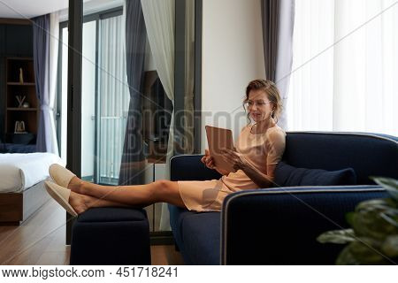 Mature Female Entrepreneur Wearing Slippers When Sitting On Couch At Hme And Reading Articles On Tab