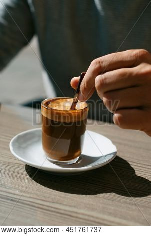 closeup of a young man stirring his machiatto, served in a glass, with a coffee spoon sitting at a table of a sidewalk cafe