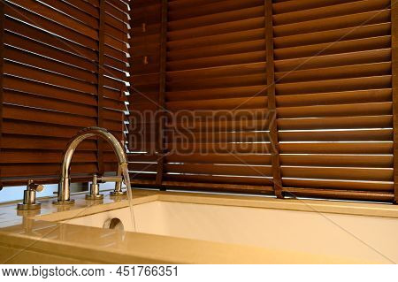 Close Up Of water Flow From Water Tap Into Bathtub With Wooden Venetian Blinds On Background In Bath