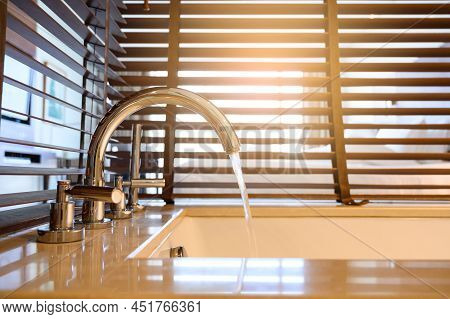 Close Up Of water Flow From Water Tap Into Bathtub With Wooden Venetian Blinds On Background In Bath