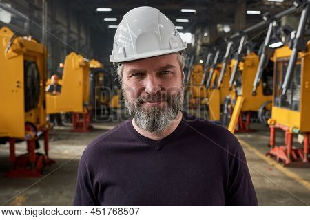 Portrait Of Mature Bearded Worker In Work Helmet Looking At Camera While Standing In Warehouse