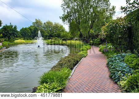A Pond Filled With Water With A Spray Jet Fountain In A Park With Pedestrian Sidewalks Made Of Stone