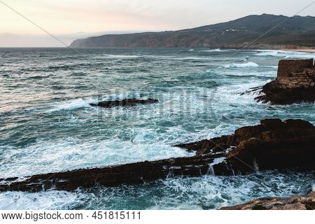 Guincho Beach At Sunset With Turquoise Waves And Foam Splashing Against Rocks In The Atlantic Ocean 