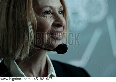 Face of happy mature businesswoman with microphone by her chin standing against interactive oard with sketches during speech