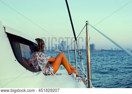 Young Woman In A Dress Sits On The Deck Of A Sailing Yacht. Back View Of Woman In Blue Dress Enjoyin