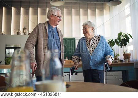 Senior Man And Woman Walking With Walker Indoors In Retirement Nursing Hme.