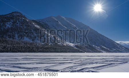 There Are Tire Tracks On The Frozen And Snow-covered Lake. At The Foot Of A Picturesque Mountain Ran