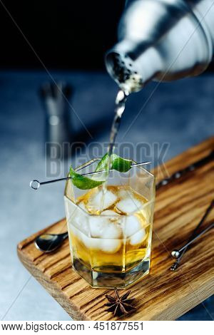 Alcohol Fresh Rum Cocktail Pouring From Shaker Into Gless With Lime Peel Ice On Wooden Desk At Black