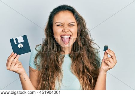 Young hispanic girl holding floppy disk and sdxc card sticking tongue out happy with funny expression. 