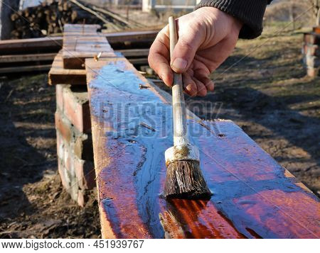 A Man's Hand With A Brush Applies Technical Brown Oil On A Long Board Outdoors In The Construction P
