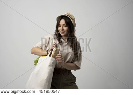 Smiling Young Woman In Beige T-shirt And A Hst With A Mesh Eco Bag Full Of Vegetables And On A White