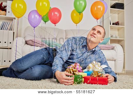 Man lies on carpet among gift boxes and birthday air balloons and looks dreamily at ceiling