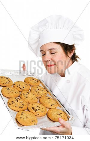 Pretty, mature female chef in her uniform, inhaling the aroma of a tray of her fresh baked chocolate chip cookies.  Isolated on white background