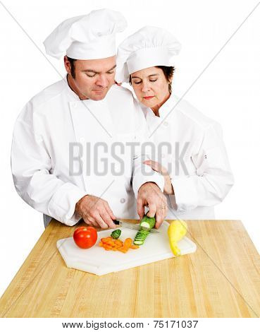 Female chef watches her male student chop vegetables on a cutting board in cooking class.  Both are in full chef's whites.  Isolated on white background.  