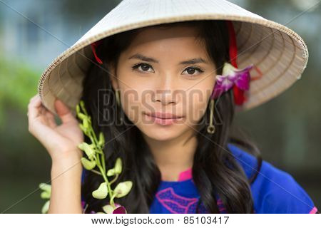 Attractive serious young Vietnamese woman in a conical straw hat with a fresh flower in her hair looking at the camera with her hand to the brim