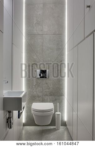 Restroom in a modern style with gray tiles and white lockers. There is a white toilet and a toilet brush, white sink with a faucet, chrome wall flush button. Backlight shines from behind. Vertical.