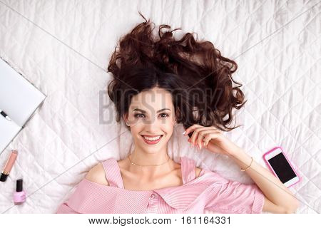 Beautiful woman lying on bed top view. Happy brunette in pink with healthy perfect hair smiling at camera. Beauty, joy, health care, fashion concept