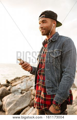 Photo of cheerful young man wearing cap walking on the beach and chatting by his phone while listening music with earphones. Looking at sea.