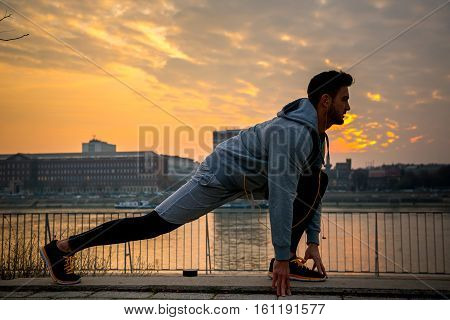 A handsome young man stretching and warming up on the riverside with the sun setting behind him
