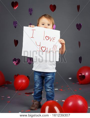 Portrait of cute adorable little white Caucasian blond boy toddler in studio with red hearts and balloons on grey background holding a piece of paper saying I love you holiday Valentine day