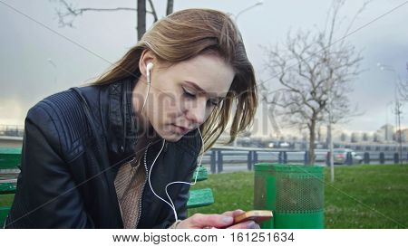 Dissapointed girl with long blonde hair in leather jacket straightens hair use gadget sitting on the bench in the wind listen headphones, close-up