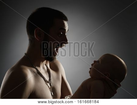 Bearded young dad with naked torso and thick silver chain over his neck with newborn in hands speaking to him affectionately