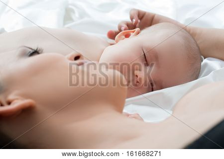 Lovely baby boy sleeping with his mother lying on white linen looking affectionately at him and gentley touching his ear close up studio shot