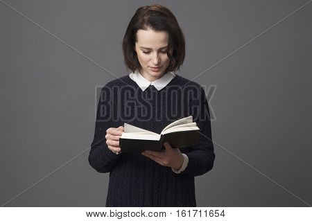 A woman in a blue sweater with a white collar reading a book gray background