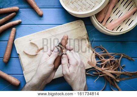 Male hands peeling an orange carrot on blue background. Concept of healthy food preparation. Peeled carrots in steamer. Vintage Retro processing Hands in frame. Top down view. Flat lay.