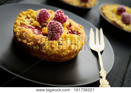 Sweet homebaked raspberry jam crumble tart with frozen raspberries on black plate on black wood boards with gold fork. Selective focus on the front raspberry. Concept of home baking.