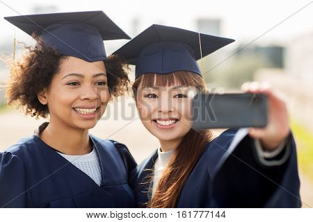 education, graduation, technology and people concept - group of happy international students in mortar boards and bachelor gowns taking selfie by smartphone outdoors