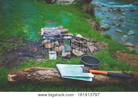 log is lying near a campfire which is near the river a map and an ax lying on a log pot mug and coffee geyser on the grass near a campfire.