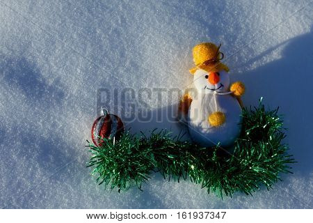 Cute happy toy snowman with green garland and red ball laying on natural white snow field and wishing Merry Christmas and a Happy New Year