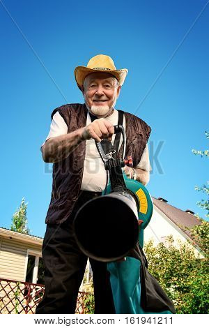 Autumn in a garden. Senior man removes fallen leaves in his garden using leaf blower.